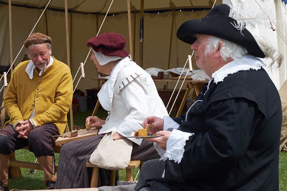 Manservant from the 1640s dressed in black and white, village couple sitting nearby.