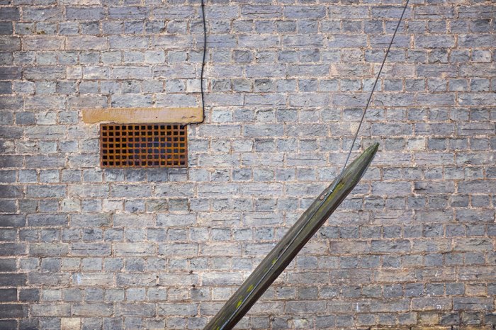 A ventilation grate, some wires or cables, a piece of wood at an angle, and everything against a brick wall.